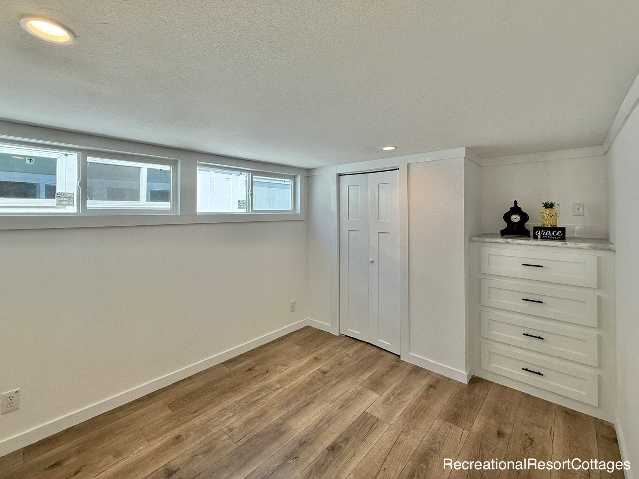 Minimalist room with light wood flooring and white walls. Small windows let in natural light. Features a closet and a set of drawers with decor.
