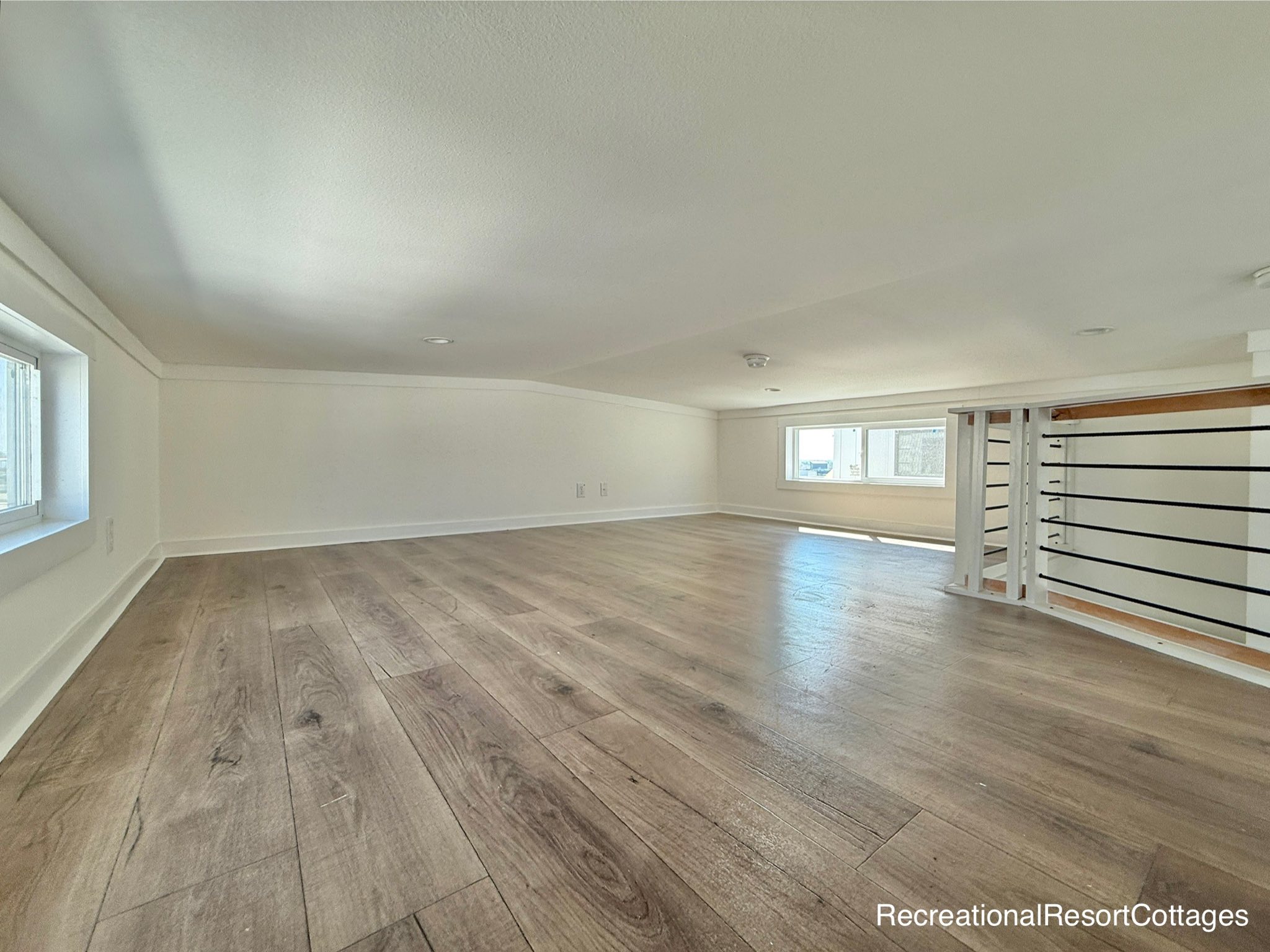 Spacious attic room with light wood flooring, white walls, and a railing leading to stairs. Natural light streams through two small windows, creating a bright and airy atmosphere.
