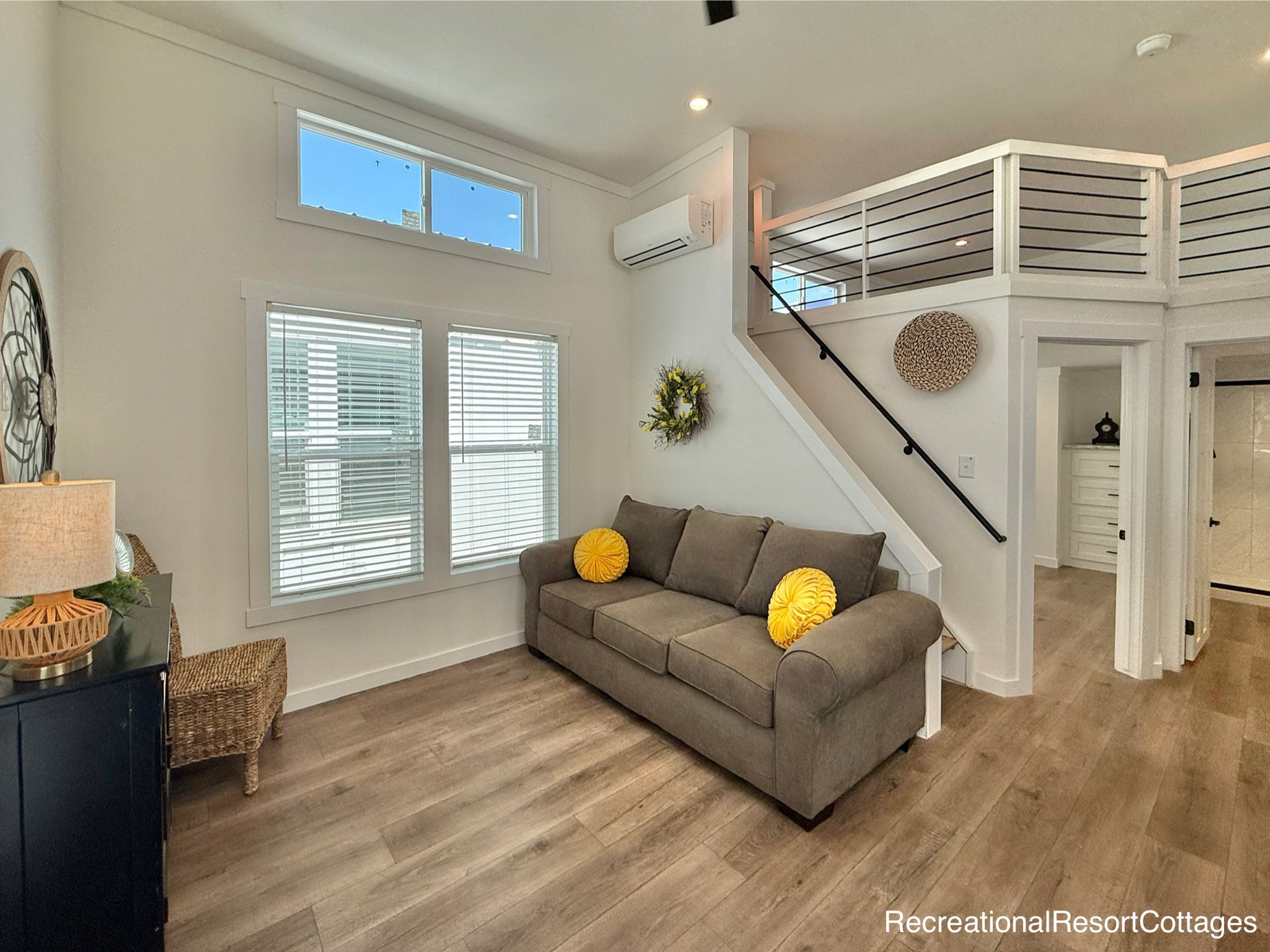 Cozy living room with a gray sofa adorned with yellow pillows, light wood floors, and large windows. A staircase leads to an upper loft area.