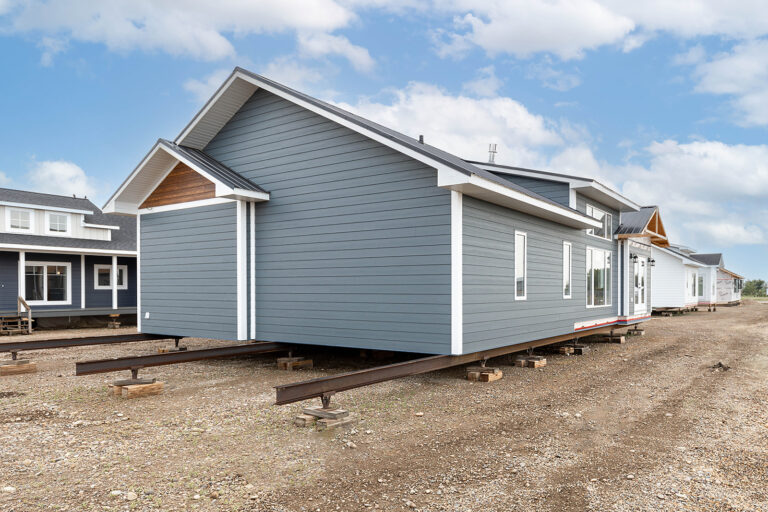 A blue modular home sits on a bare, gravelly ground, supported by metal beams. Other similar homes are visible in the background under a cloudy sky.