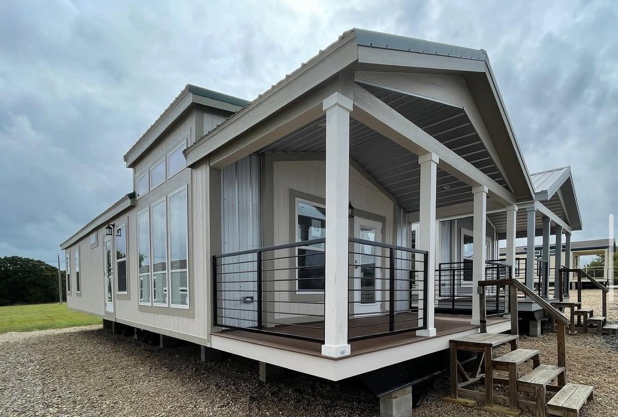 Modern tiny house with a pitched roof and large windows. It features a wooden porch with a railing and steps, set under a cloudy sky.
