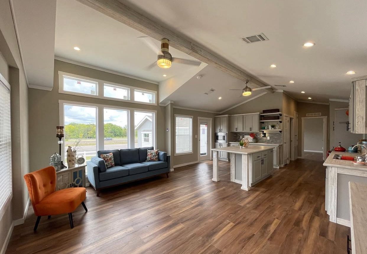 Spacious open-plan living room and kitchen with wood floors. Features orange chair, blue sofa, large windows, and modern kitchen island. Bright and inviting.