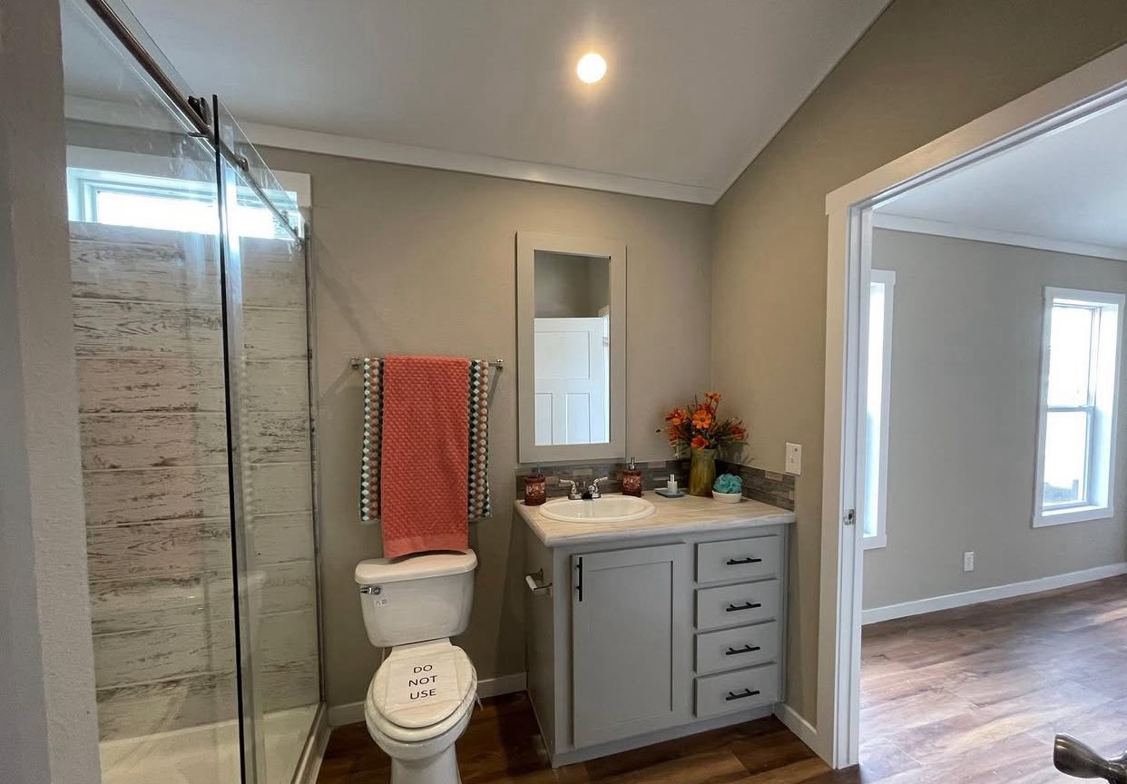 Modern bathroom with a glass-enclosed shower, a toilet labeled "Do Not Use," and a vanity with an orange towel and floral decor, adjacent to a bright, wood-floored room.