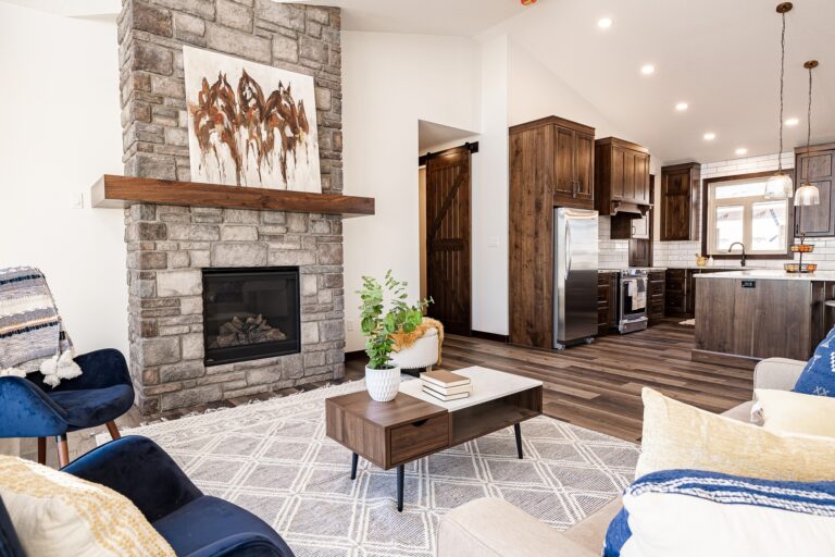 A cozy living room with a stone fireplace, abstract art, and wooden accents. A modern kitchen is visible. Soft lighting creates a warm, inviting ambiance.