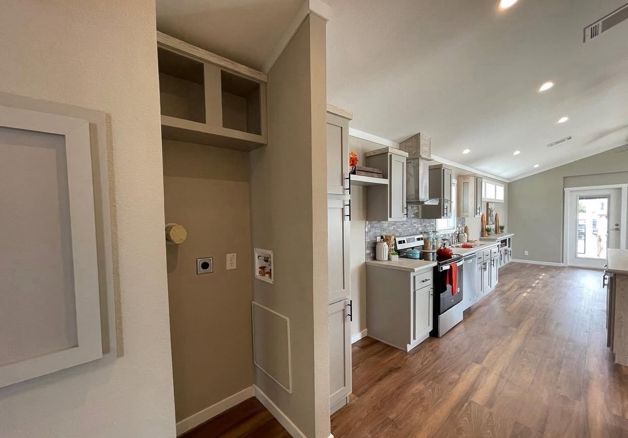Modern kitchen with light gray cabinets, stainless steel appliances, and wood flooring. Open layout leads to a door with large windows, creating a bright, airy feel.