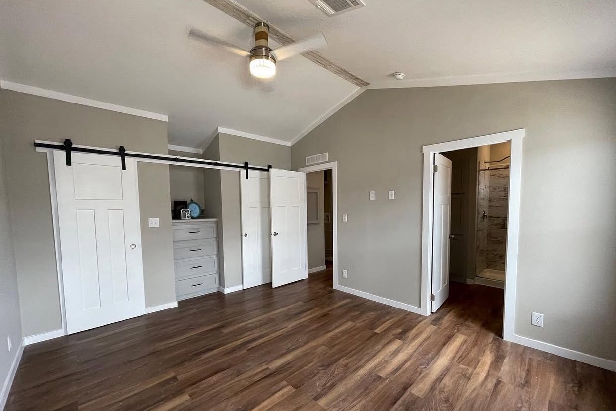 Spacious room with light gray walls and wood flooring, featuring a ceiling fan. Double sliding doors reveal a closet with shelves. A door on the right leads to a bathroom.