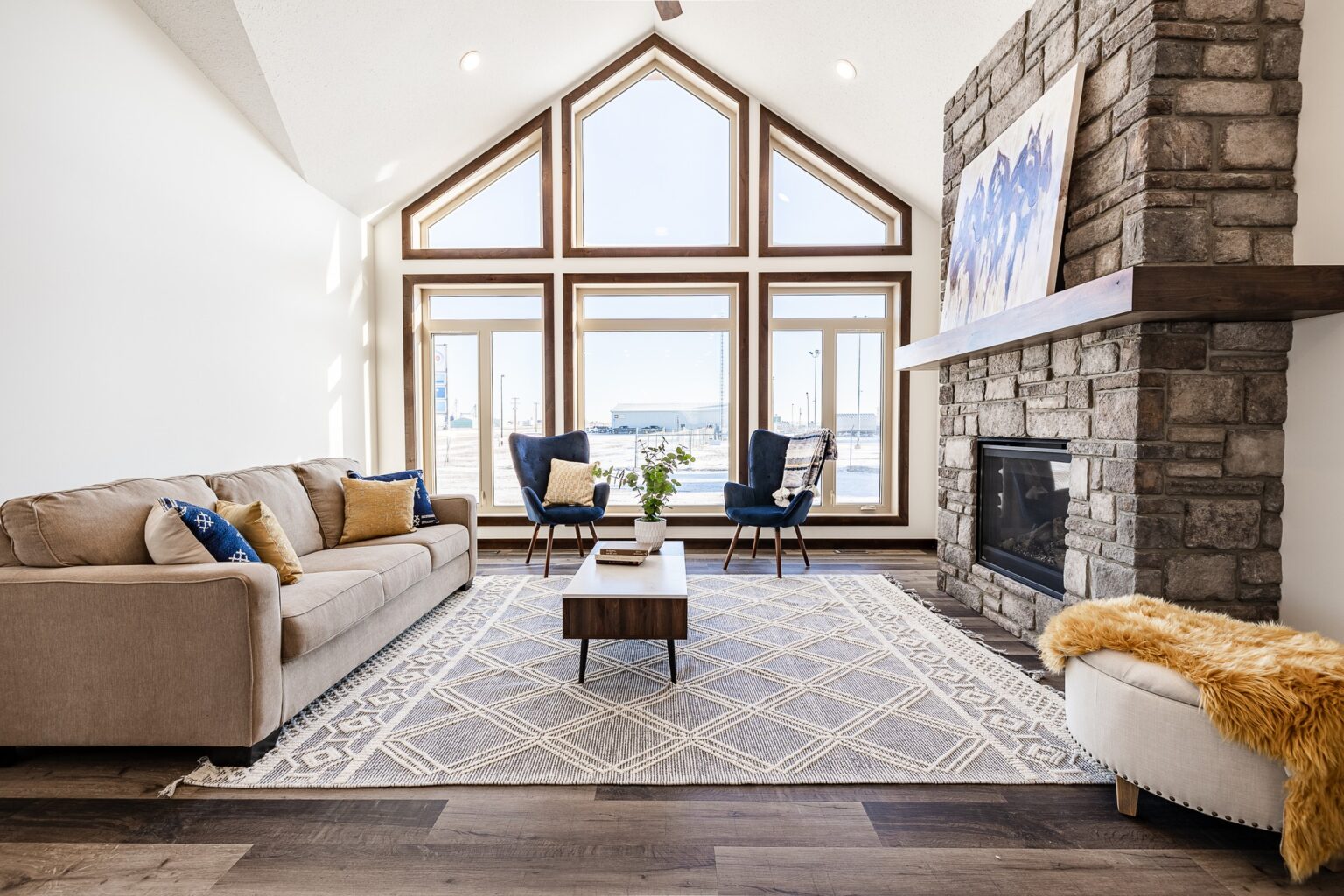 A spacious, bright living room with large triangular windows, beige sofa, two blue chairs, stone fireplace, and geometric rug, exuding warmth.