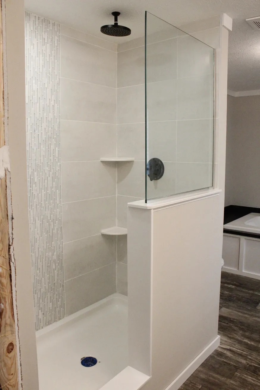 Modern shower with light gray tiles, vertical mosaic accent strip, and dual corner shelves. Glass partition and dark showerhead. Clean, sleek design.