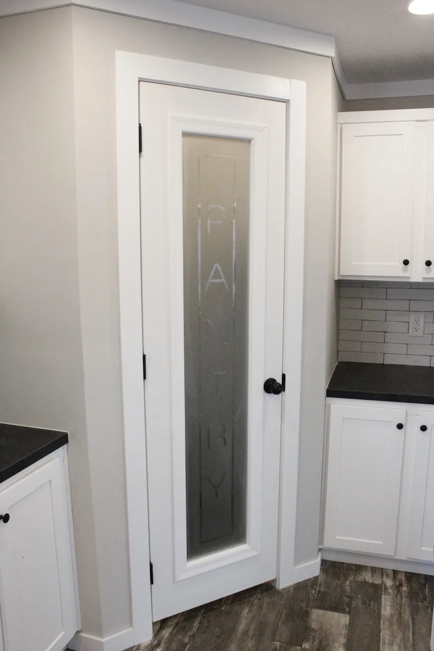 White kitchen with a frosted glass pantry door labeled "Pantry" and dark countertops. The space is modern and clean, with light gray walls.