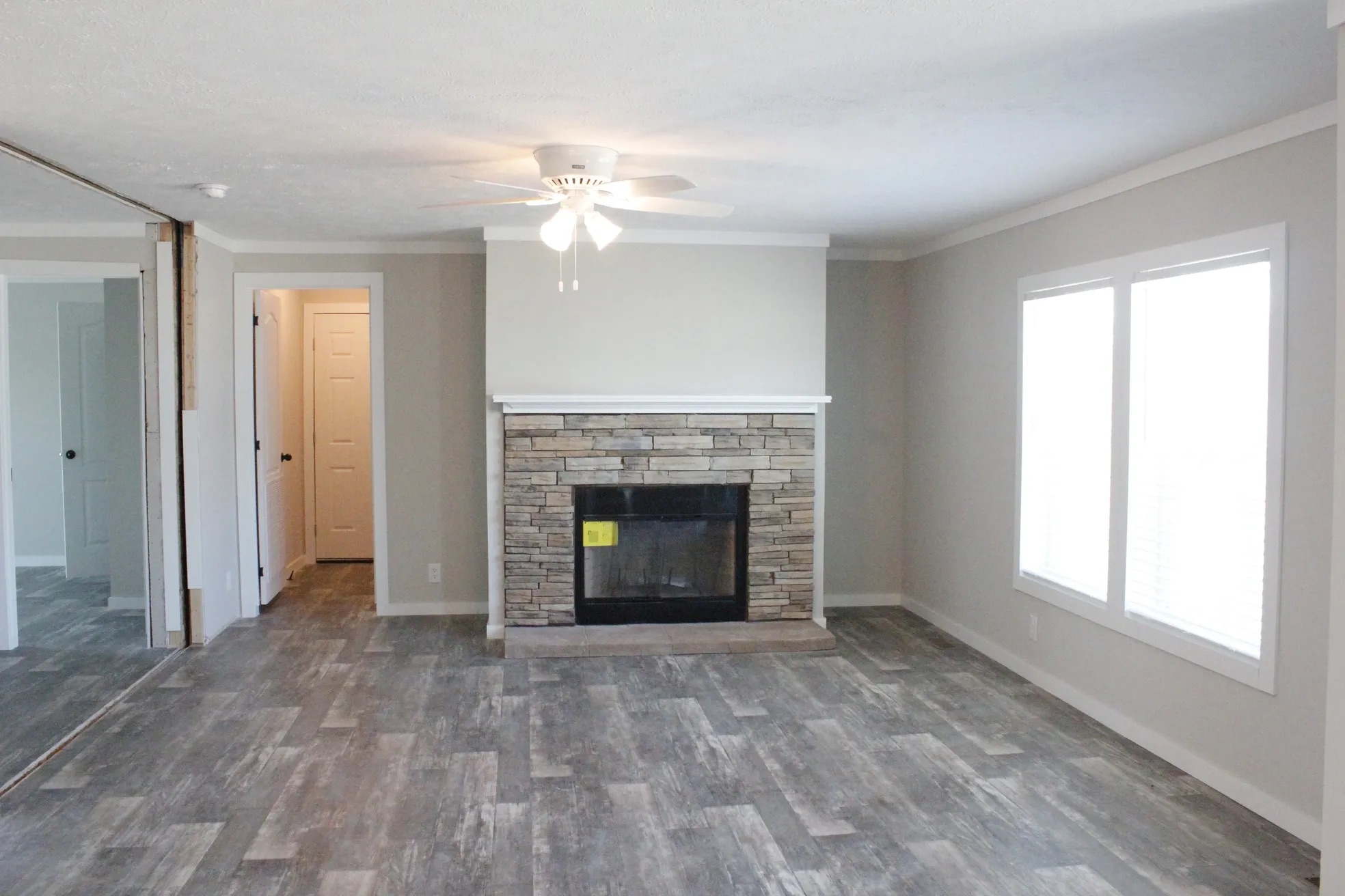 A spacious living room with gray tile flooring, a stone fireplace, and two large windows. A ceiling fan is mounted on the white ceiling.