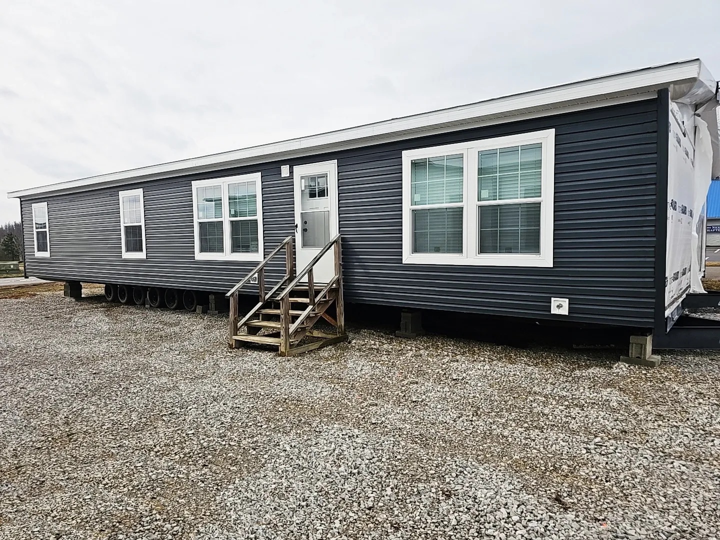 A modern, dark-colored mobile home with multiple large windows sits on a gravel plot. Wooden steps lead to the door, suggesting a simple yet functional design.