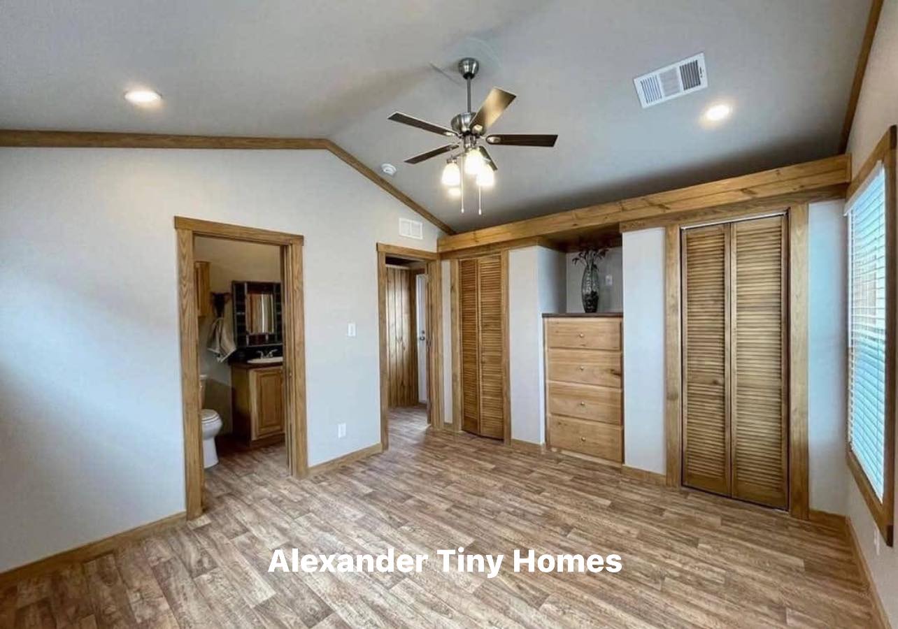 Spacious tiny home interior with wooden floors and ceiling fan. Features two doorways, closets, natural light, and subtle warmth. "Alexander Tiny Homes" text.