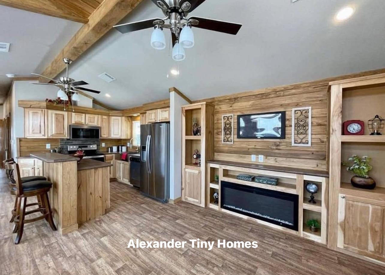 Modern tiny home kitchen with natural wood cabinets, stainless steel appliances, and a cozy living area featuring a faux fireplace and built-in shelves.