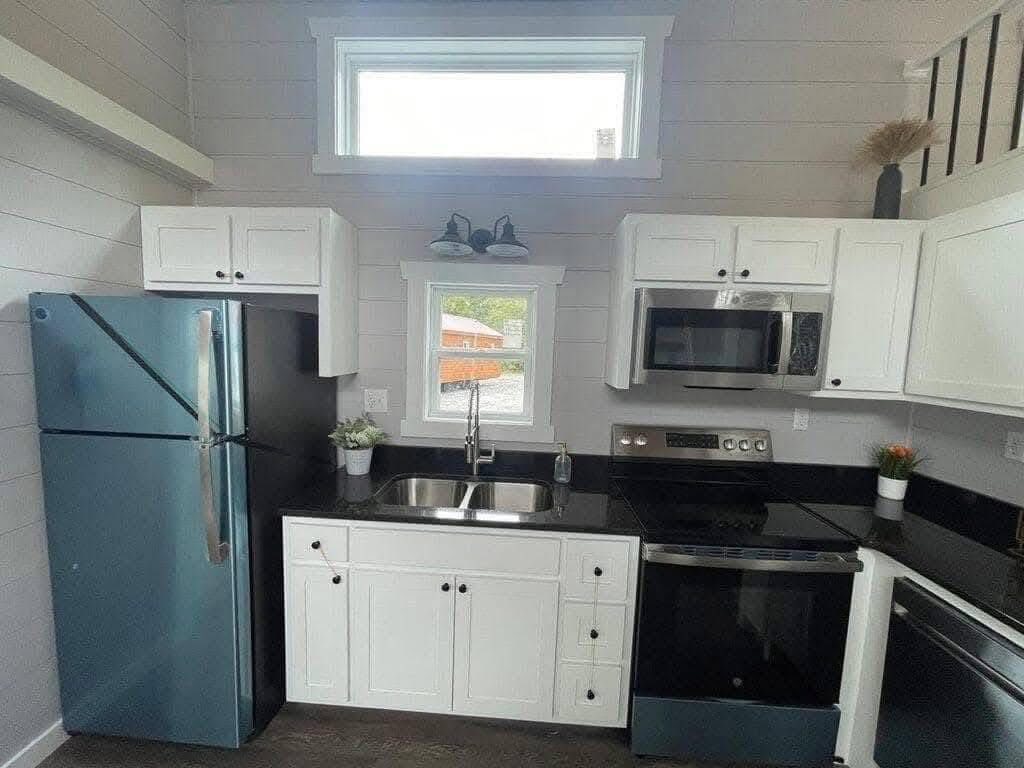A compact modern kitchen with black countertops, white cabinets, double sink under a window, stainless steel fridge, oven, and microwave, conveying a sleek, clean ambiance.