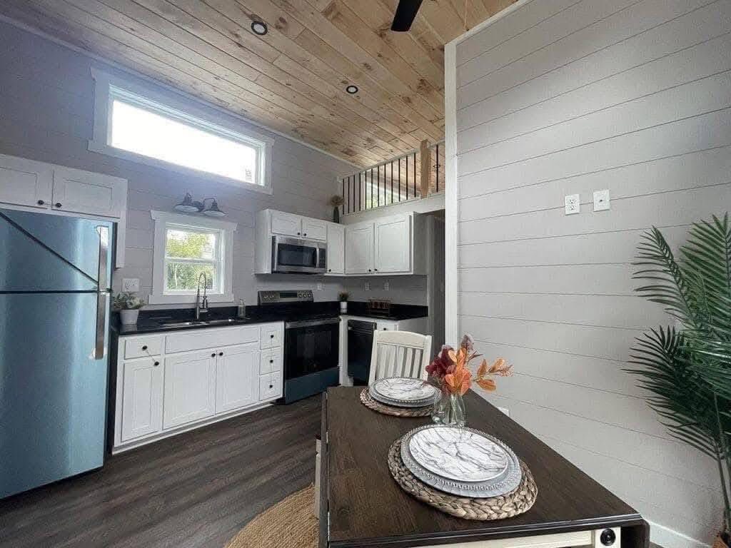 Bright, cozy kitchen with white cabinets, stainless steel appliances, and rustic wood ceiling. A small dining area with plates and autumn flowers adds warmth.