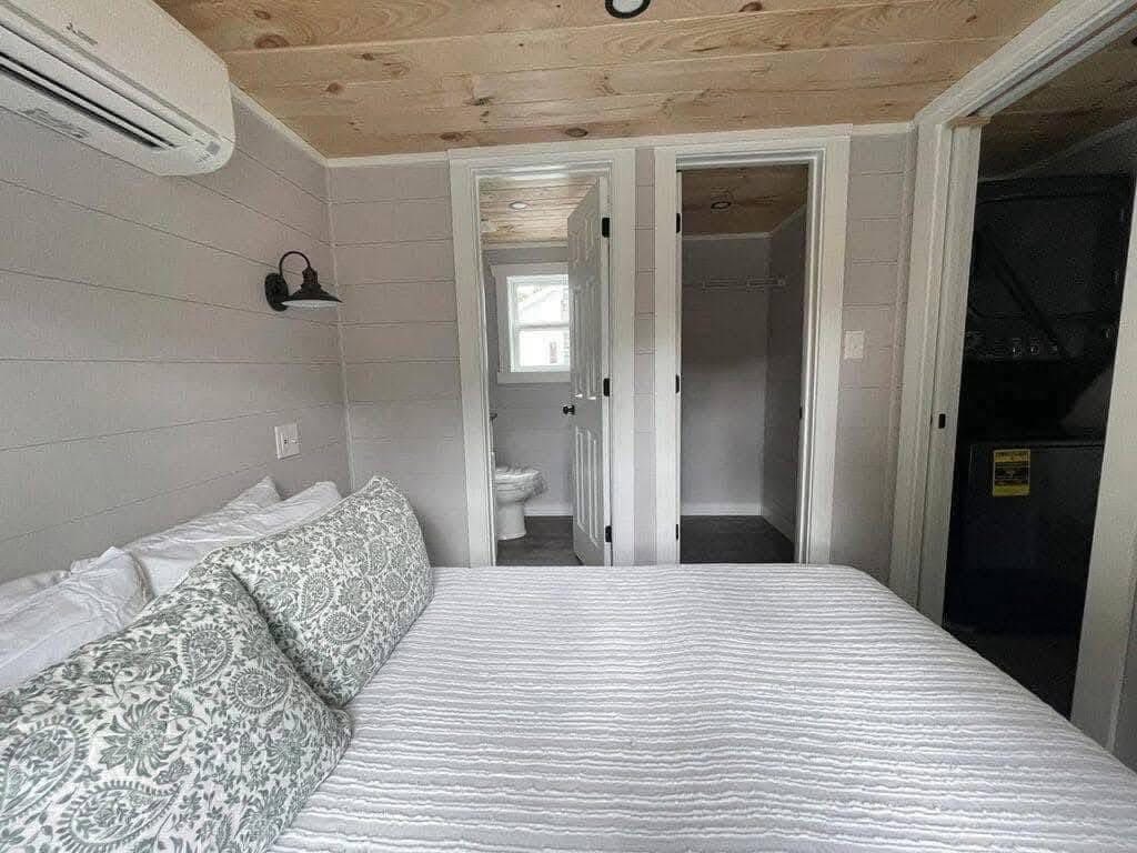 Cozy bedroom with light gray walls, wood ceiling, and a striped bedspread. Floral pillows, a vintage wall lamp, and a door to a bathroom create a serene feel.