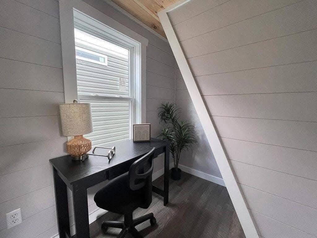 A small, cozy home office with a black desk, swivel chair, lamp, and radio near a window. A potted plant adds greenery. Light, neutral tones create a calming vibe.