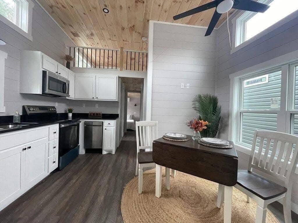Compact kitchen and dining area in a tiny home with white cabinetry, dark countertops, and wooden accents. Cozy, modern, and efficient atmosphere.