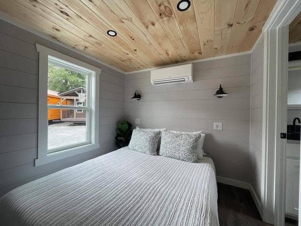 Cozy bedroom with gray walls, wooden ceiling, and a large window. A bed with white bedding and pillows is centered, with wall sconces and an air conditioner above.
