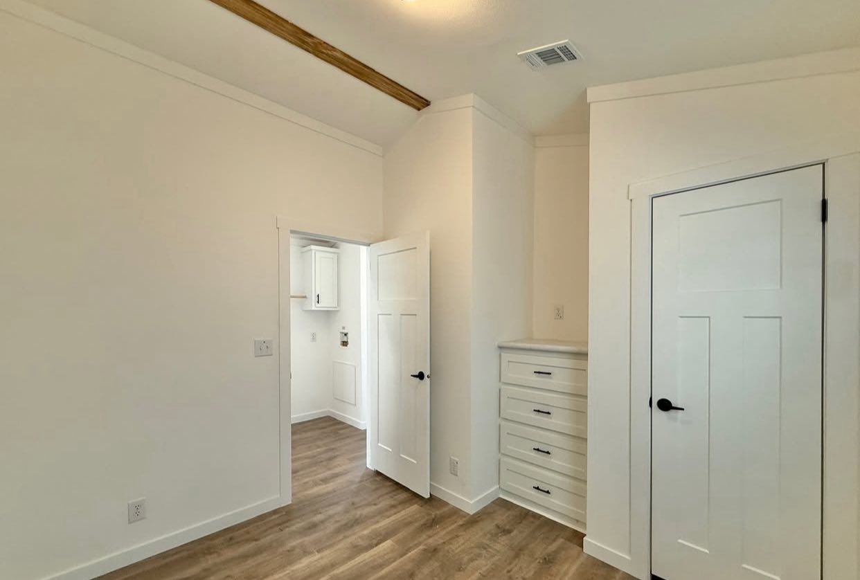 Small, bright room with wood flooring, white walls, and wood beam on ceiling. It features an open door, a closet door, and built-in drawers.