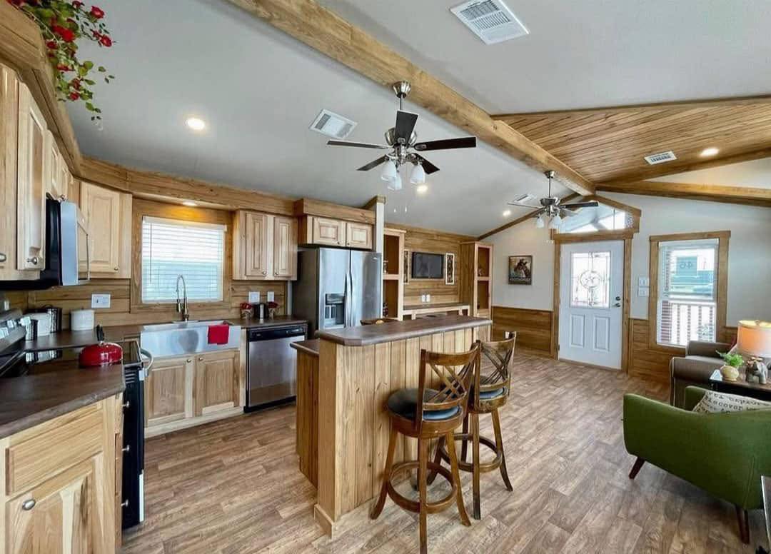 Spacious kitchen and living area with wood accents and flooring. Features include a central island, stainless steel appliances, and cozy seating. Warm, inviting atmosphere.