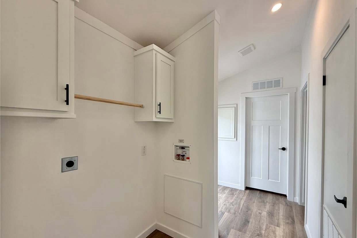 Laundry room with white walls and cabinets. A wooden rod is mounted between cabinets on the left. Wood flooring and a door lead to an adjacent room.