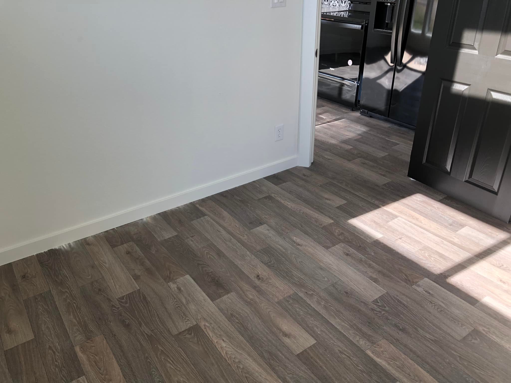 Sunlit room with light wood laminate flooring. A black door is ajar, leading to a kitchen area with dark appliances, creating a modern feel.