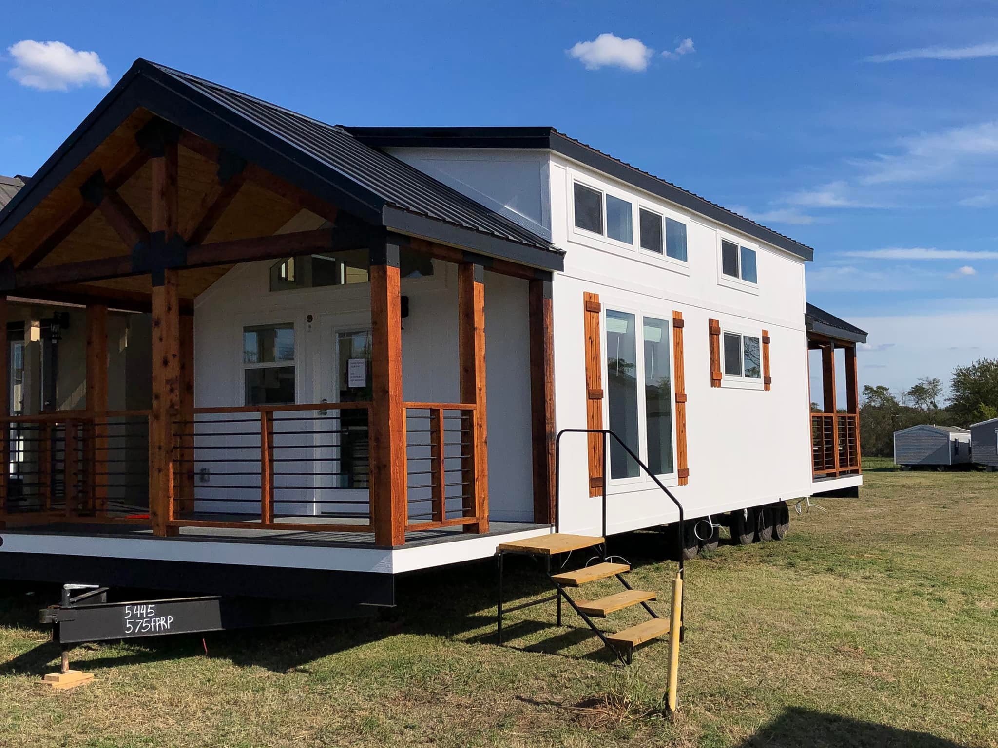 A modern tiny home with white siding and wooden accents sits on a grassy field under a clear blue sky. A small porch is visible with railings.