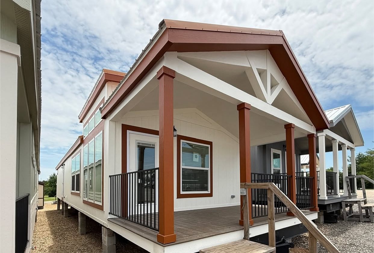 A small, modern prefab house with white walls and red-brown trim features a front porch with black railings, set against a cloudy sky backdrop.