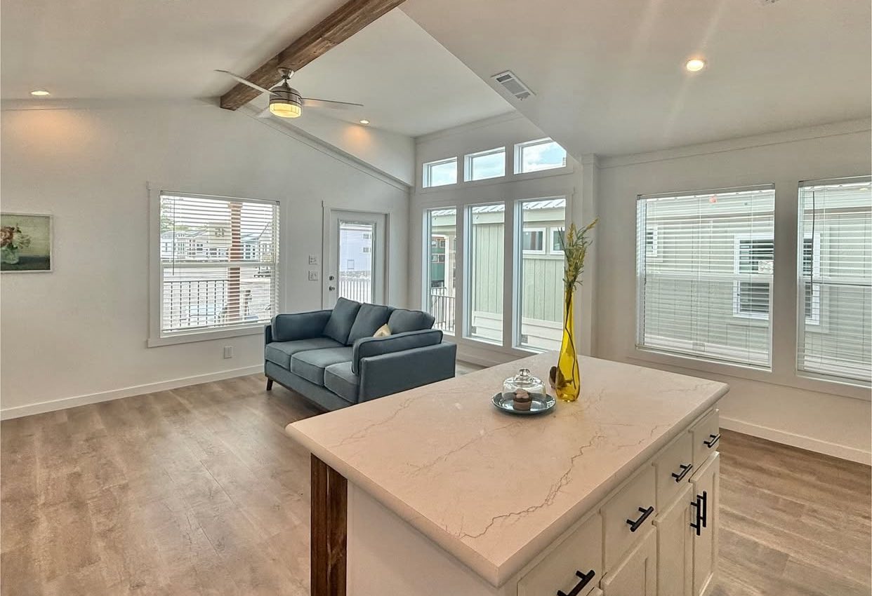 Bright, modern living room with light wood flooring. A blue sofa sits near tall windows, and a white island with a yellow vase adds elegance. Cozy and airy ambiance.