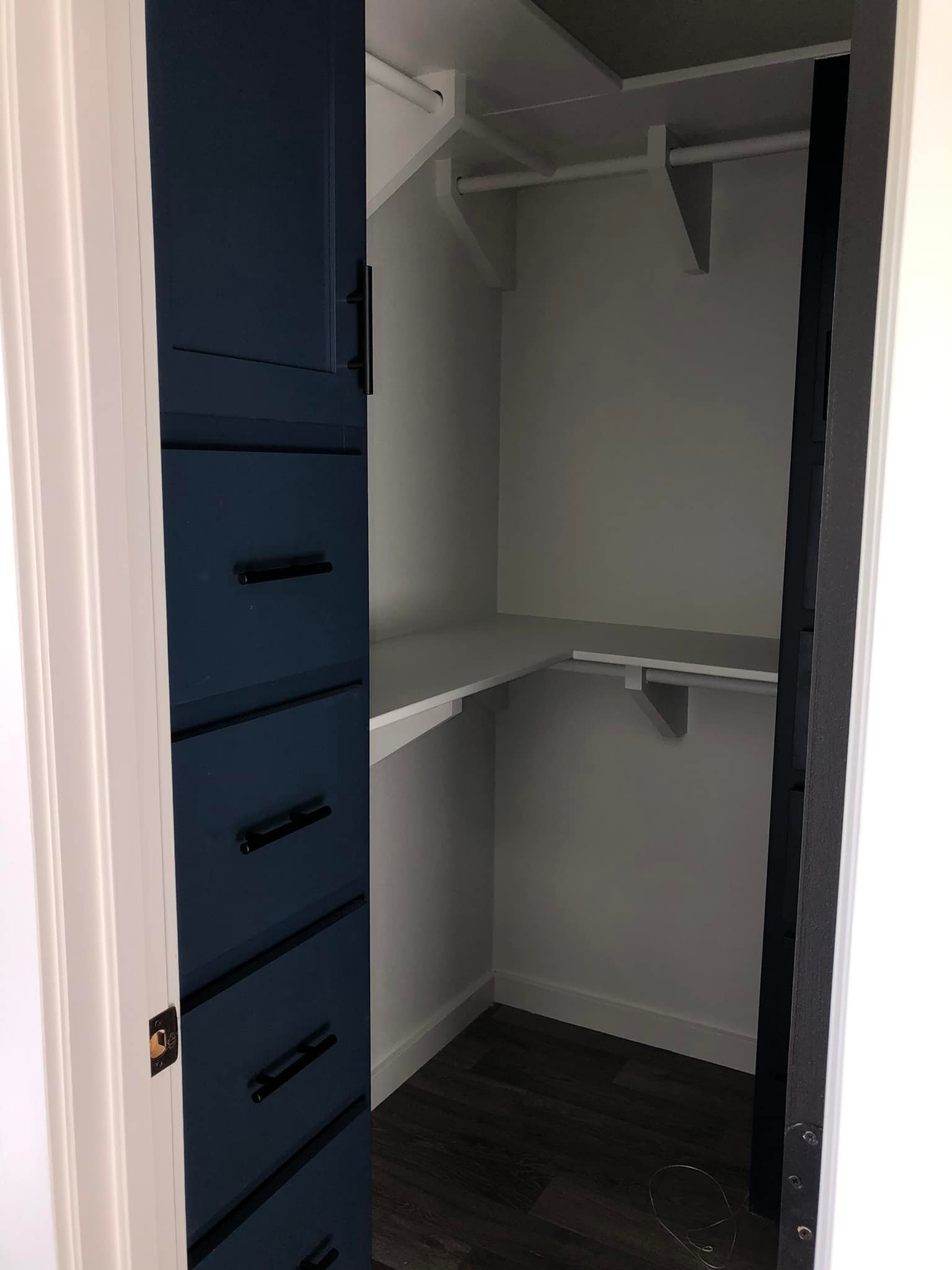 A small gray closet with light wood flooring features a navy cabinet with black handles on the left and white shelves and rods for hanging clothes.