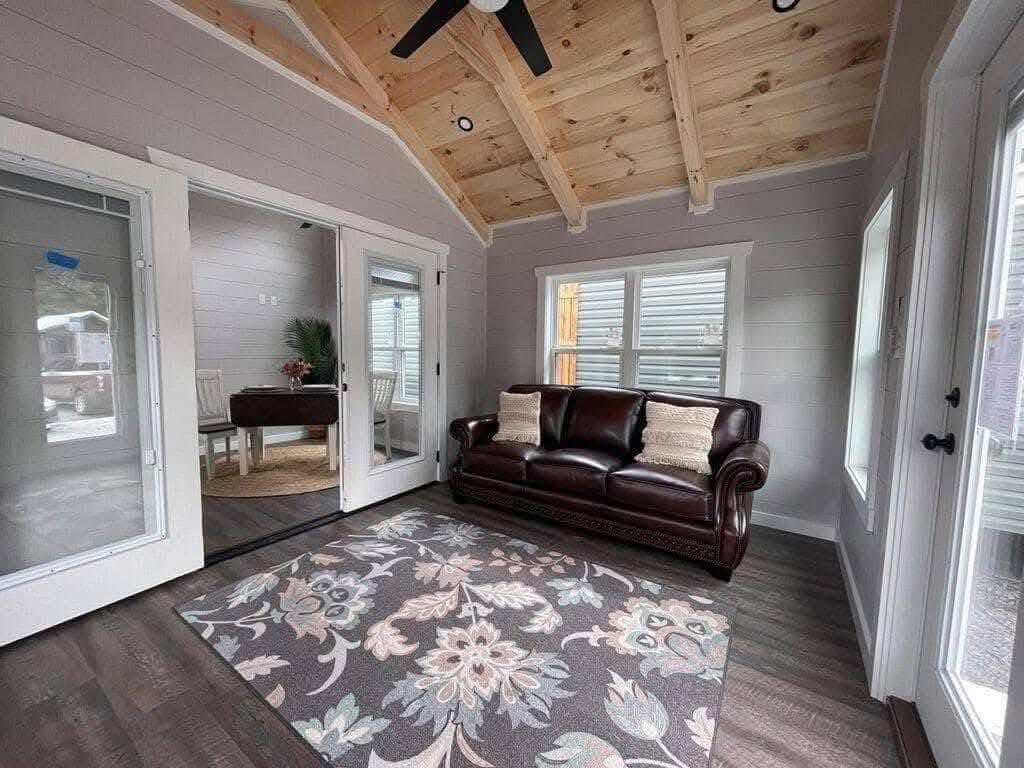Cozy room with vaulted wood ceiling and a dark brown leather sofa adorned with pillows. Light gray walls, floral rug, and glass doors enhance the welcoming ambiance.