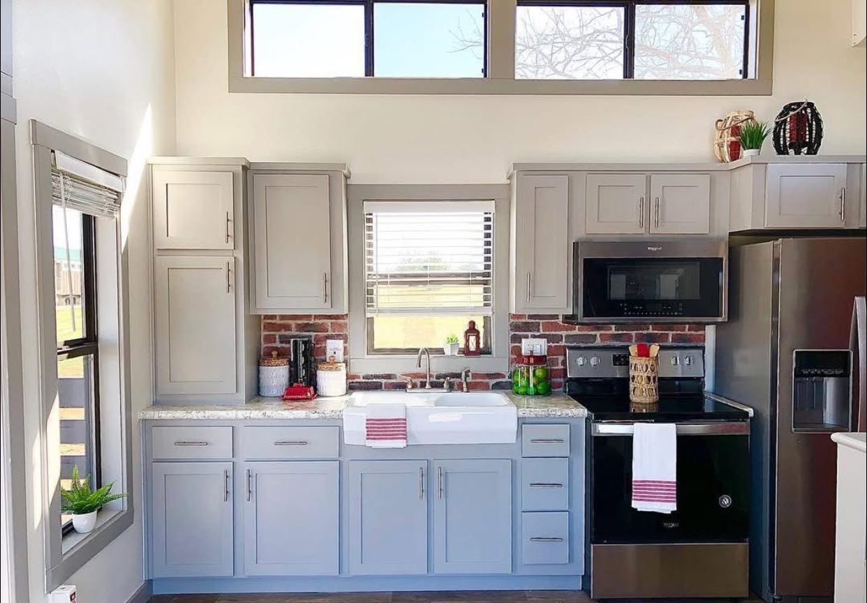 Bright kitchen with light gray cabinets, stainless steel appliances, and a white farmhouse sink. Brick backsplash adds rustic charm. Ample natural light fills the space.