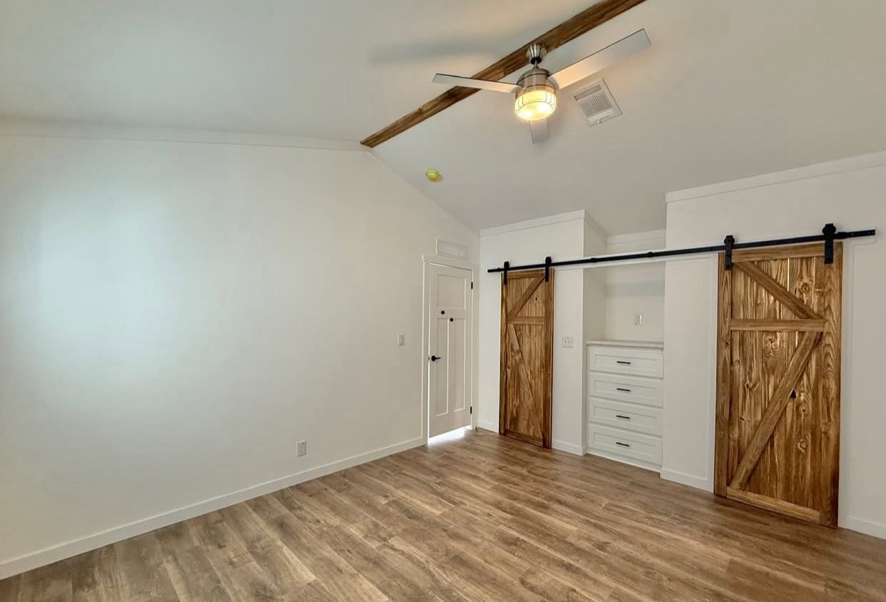 Minimalist room with white walls and wood flooring. Features rustic wood sliding doors on a closet, modern ceiling fan, and bright, airy atmosphere.
