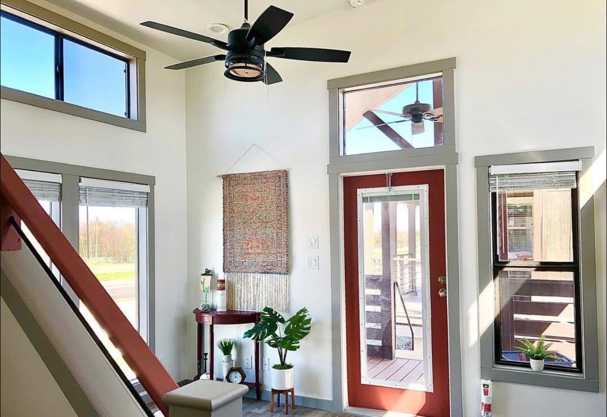 Bright and airy room with large windows, a red-framed glass door, and a ceiling fan. Decor includes plants, a woven wall hanging, and minimalist furniture.
