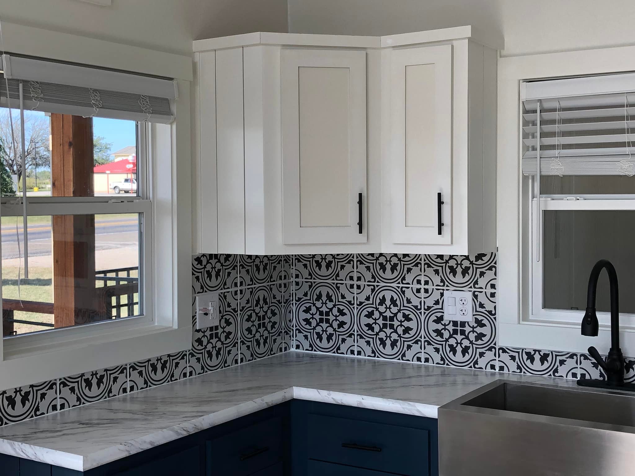 Modern kitchen corner with white cabinets, black handles, patterned tile backsplash, marble countertop, dark blue lower cabinets, and a large sink.