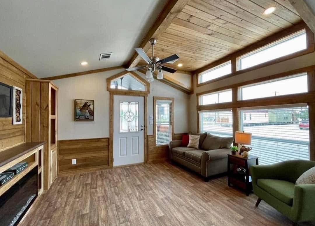 A cozy living room with rustic wood paneling and ceiling. It features a ceiling fan, windows with natural light, a sofa, and an armchair. Warm and inviting ambiance.
