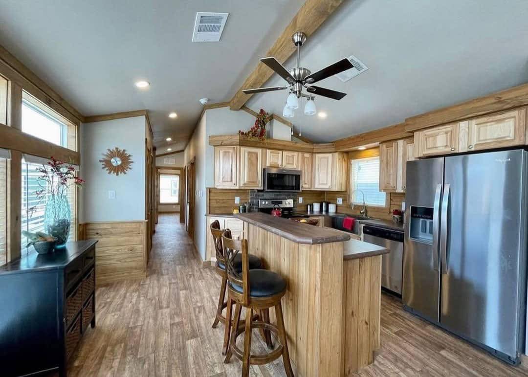 A cozy kitchen with wooden cabinets, a stainless steel fridge, and a bar counter with two chairs. Warm lighting creates a welcoming ambiance.