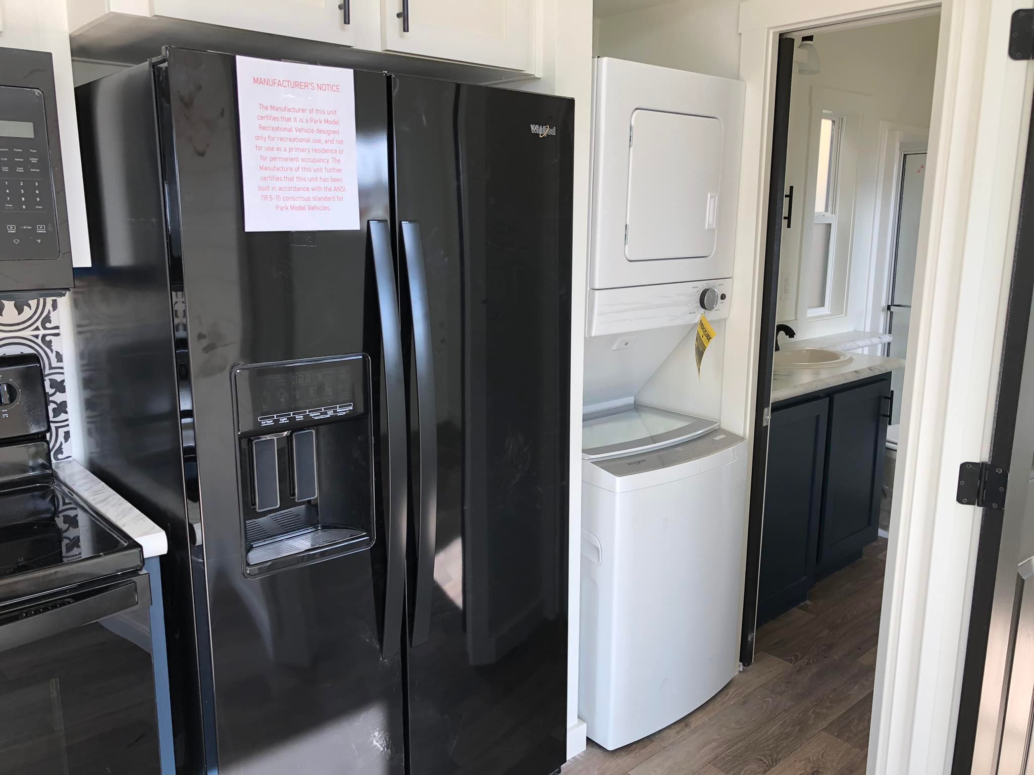 Modern kitchen with a black refrigerator next to a stacked white washer-dryer unit. A narrow hallway leads to a bathroom with dark cabinetry.