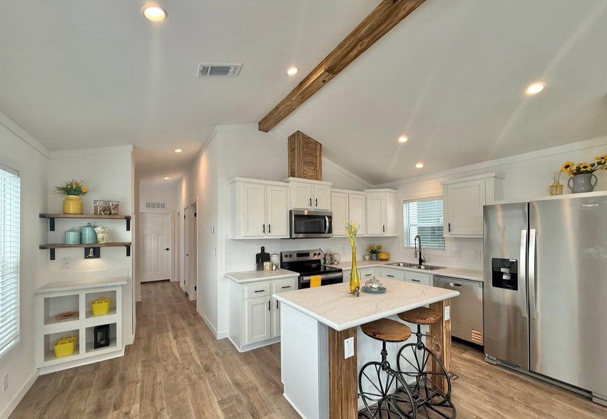 Modern kitchen with white cabinets, stainless steel appliances, and a wooden beam ceiling. A center island has bicycle-style stools, and the space feels bright and inviting.