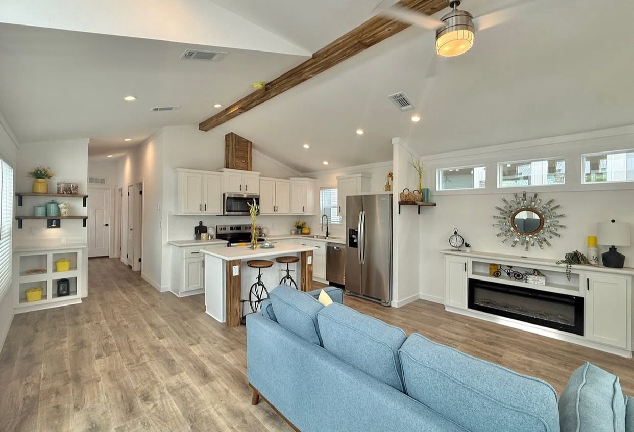 Spacious open-plan living room and kitchen with wood floors. White cabinets, stainless steel appliances, light blue couch, rustic beams, and decorative shelves.