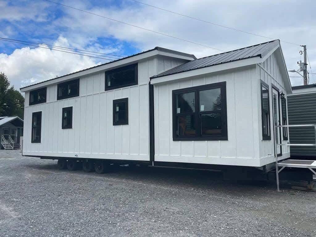 Modern white tiny house on wheels, featuring multiple large black-framed windows. It sits on a gravel lot under a partly cloudy sky.