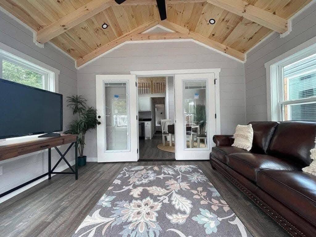 A cozy room with light gray walls and a wood-paneled ceiling. It features a dark leather sofa, patterned rug, TV, and glass doors leading to a kitchen.