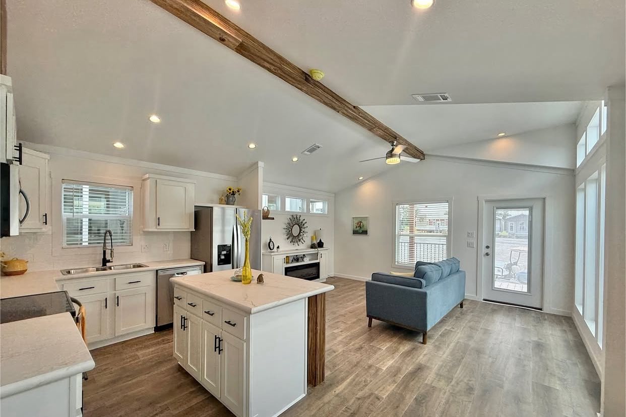 Modern open-plan living room and kitchen with vaulted ceiling and exposed beam. Features white cabinets, a center island, and a blue sofa on wood floors.
