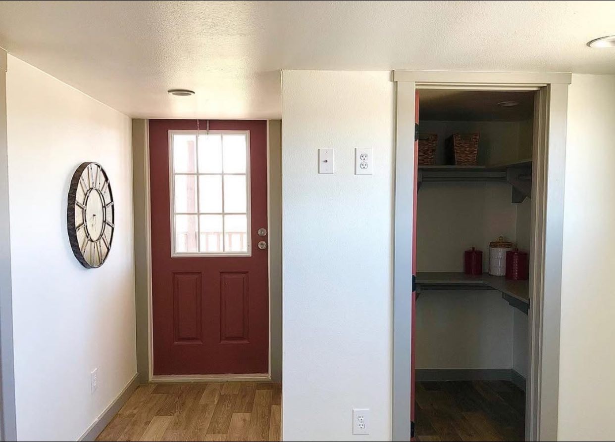 A small hallway with a red door featuring glass panels, a large wall clock, and a walk-in pantry. The space is minimalist and bright.