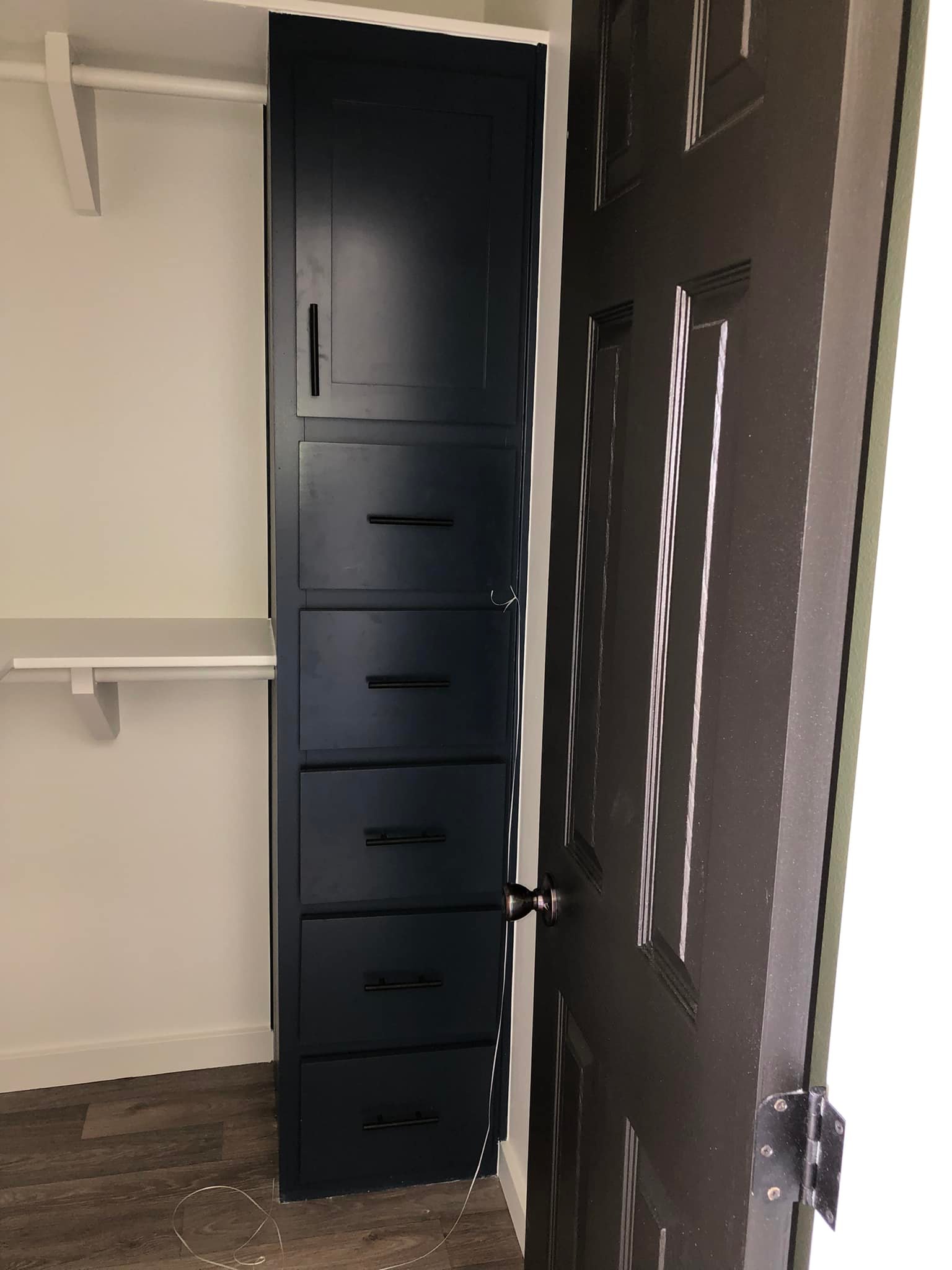 A narrow closet with dark blue shelving and drawers is visible behind a partially open brown door. The floor is wooden, and the walls are white.
