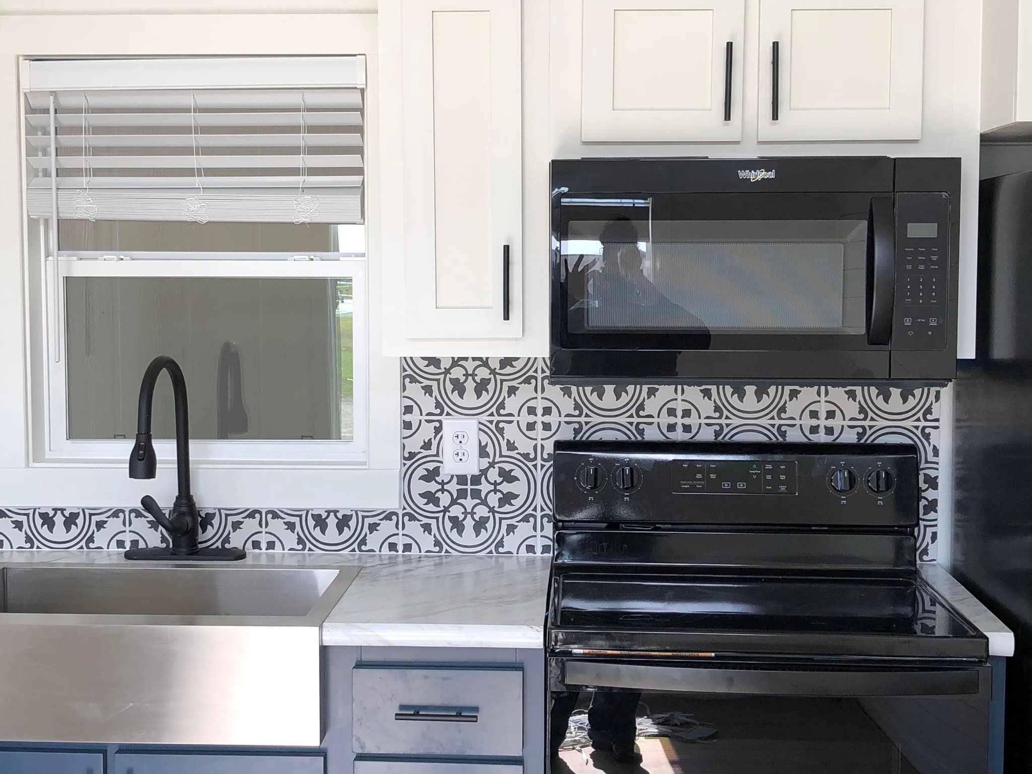 Modern kitchen with white cabinets, black handles, black appliances, and a sink. Intricate black-and-white tile backsplash adds elegance.
