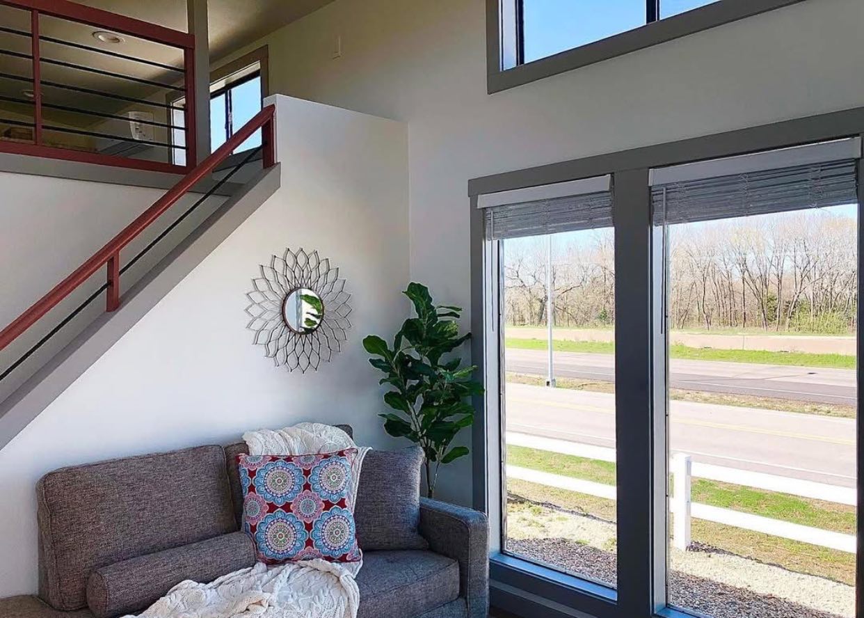 Modern living room with gray couch adorned with patterned cushions and a throw. Sunlit windows offer views of a road and greenery, with a round mirror and a potted plant nearby.