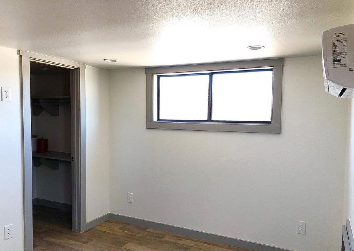 Small, minimalistic room with light-colored walls, a wood floor, and a rectangular window. Doorway leads to a closet. Air conditioning unit on the wall.