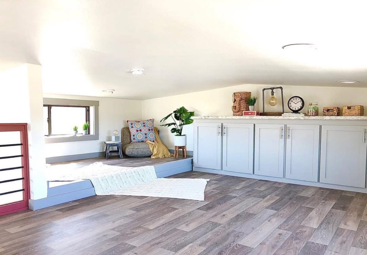 Bright, cozy loft space with light wood flooring and a large gray cabinet. A round chair with colorful pillows is in the corner, next to a window.